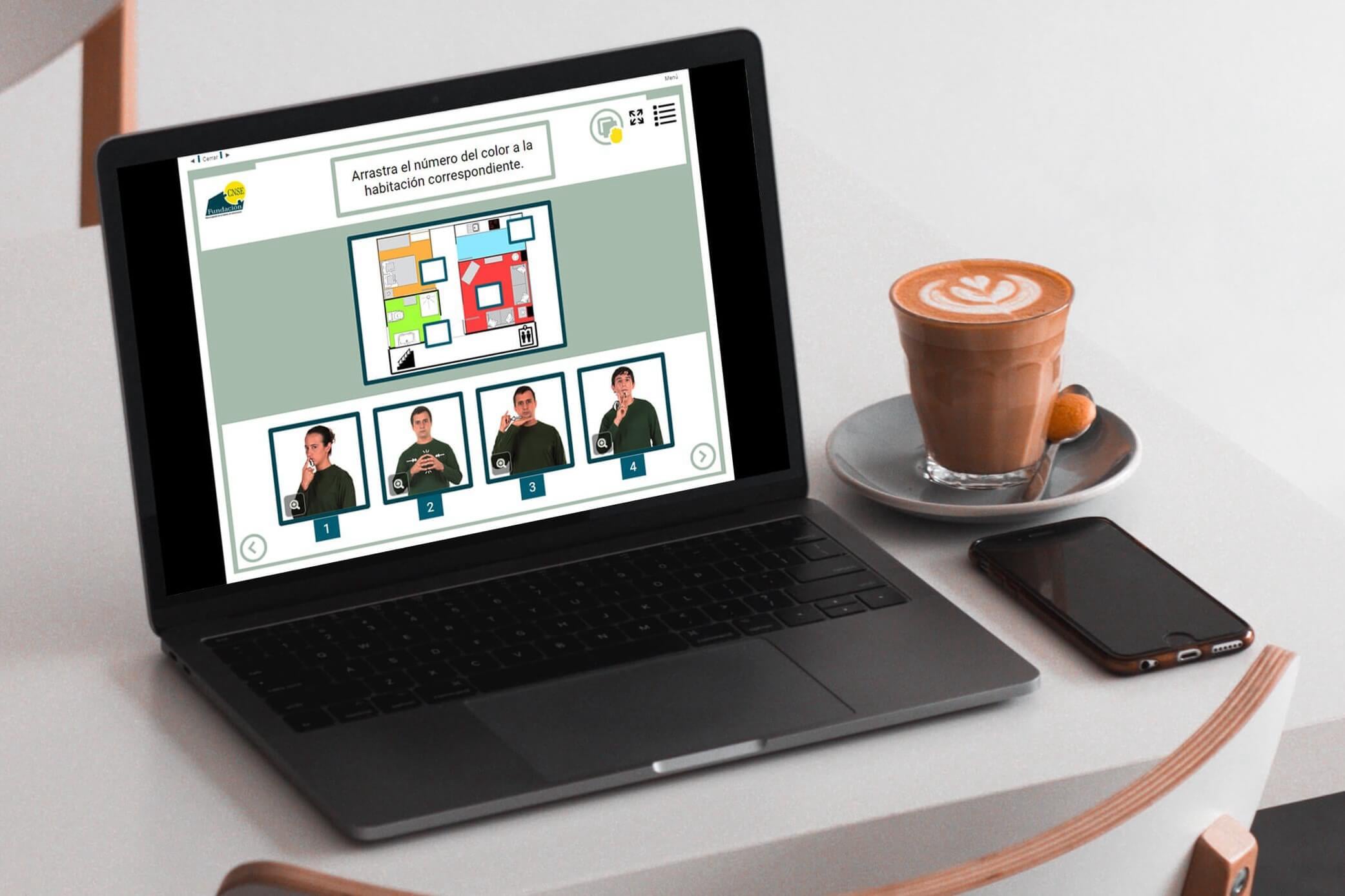 Imagen de un ordenador portátil con el curso online de lengua de signos. En la mesa hay un café y un teléfono móvil